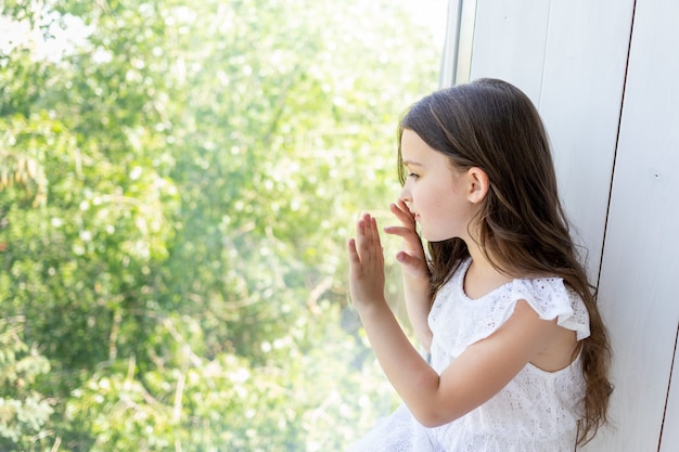 Retrato de una encantadora niña de 5-6 años mirando por la ventana