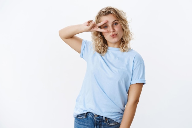 Retrato de una encantadora rubia europea divirtiéndose y disfrutando de la vida mostrando la paz o el signo de la victoria sobre los labios plegados de los ojos tontamente despreocupada y alegre mientras posa relajada sobre fondo blanco