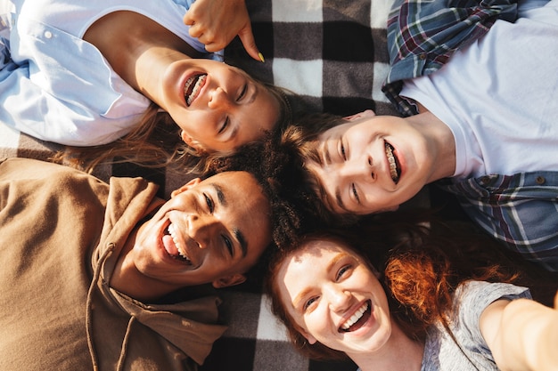 Retrato de encantadores jóvenes amigos chicos y chicas riendo y acostado sobre una manta en círculo al aire libre