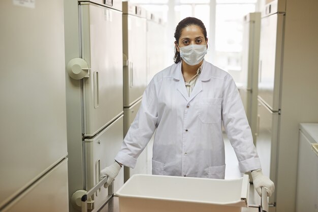 Retrato de enfermera con máscara y bata blanca mientras está de pie en el laboratorio entre neveras con muestras