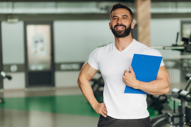 Foto retrato de entrenador personal sosteniendo portapapeles con plan de entrenamiento en el gimnasio