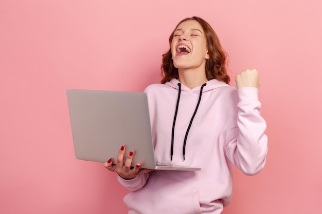 Retrato de una entusiasta adolescente de pelo rizado con capucha gritando de alegría y sosteniendo una laptop, regocijándose de la victoria. Disparo de estudio interior aislado sobre fondo rosa
