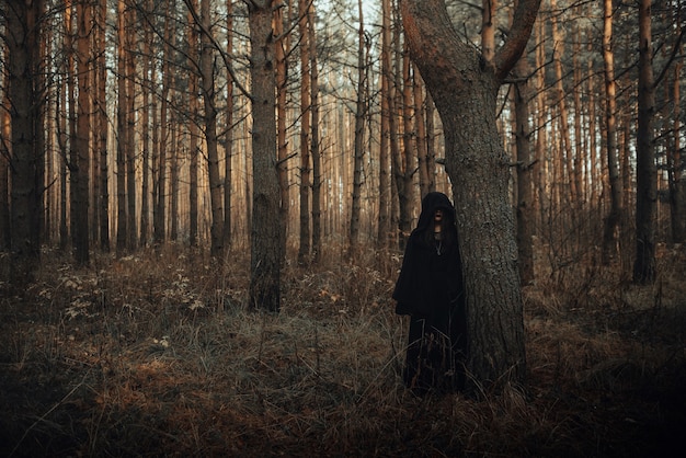 Retrato escuro de uma bruxa terrível em um terno preto na floresta perto de uma árvore