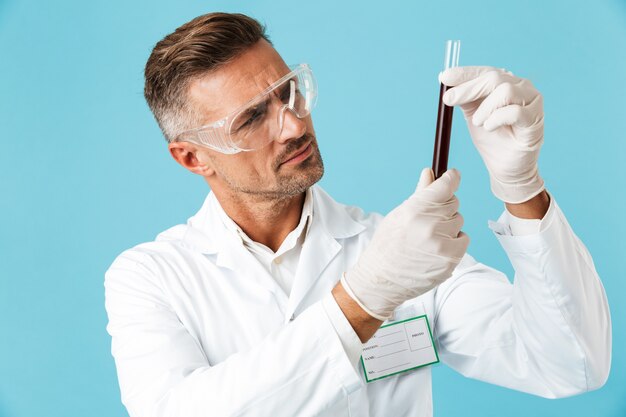 Retrato de especialista médico serio con gafas sosteniendo el tubo de ensayo con sangre, que se encuentran aisladas sobre la pared azul