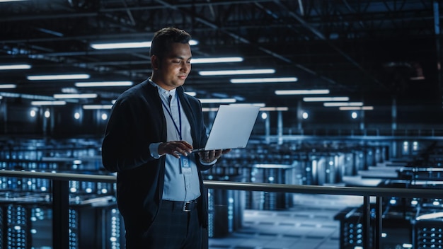 Retrato de un especialista en TI que utiliza una computadora portátil en una instalación de computación en la nube de la granja de servidores del centro de datos con un administrador de sistemas masculino que trabaja en seguridad cibernética y protección de redes