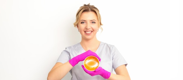 Retrato de una esteticista caucásica sosteniendo un frasco de pasta de azúcar para azucarar usando guantes rosas sobre fondo blanco