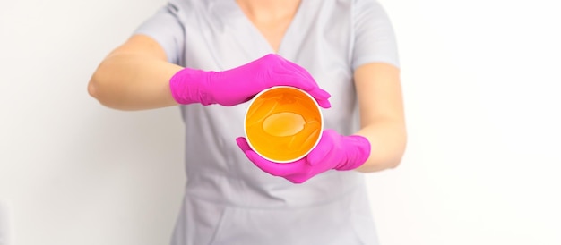 Retrato de una esteticista caucásica sosteniendo un frasco de pasta de azúcar para azucarar usando guantes rosas sobre fondo blanco