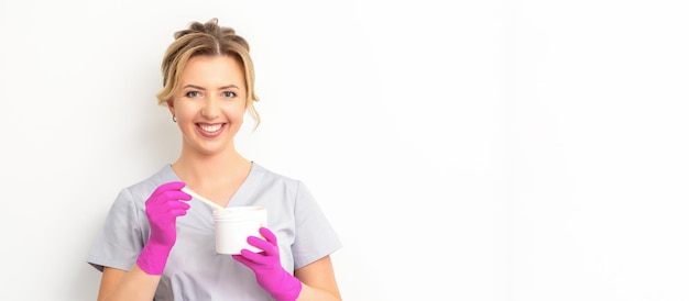 Retrato de una esteticista caucásica sosteniendo un frasco de pasta de azúcar para azucarar usando guantes rosas sobre fondo blanco