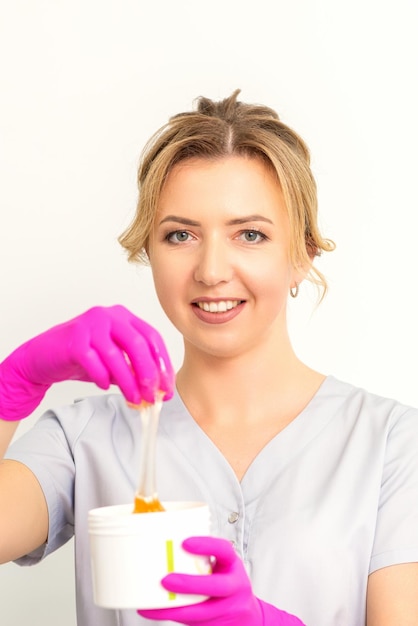 Retrato de una esteticista caucásica sosteniendo un frasco de pasta de azúcar para azucarar usando guantes rosas sobre fondo blanco