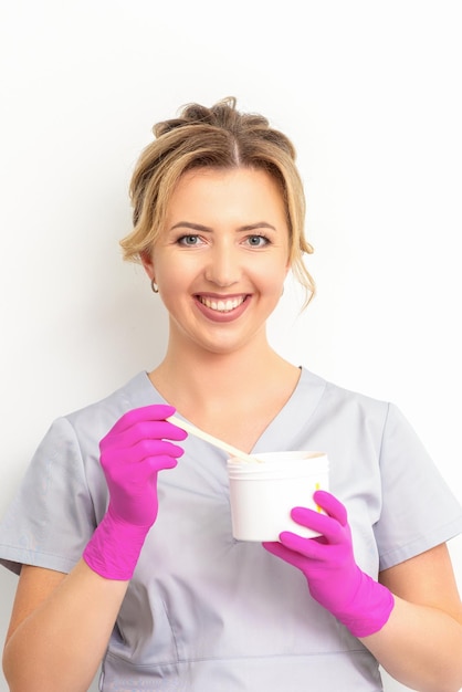 Retrato de una esteticista caucásica sosteniendo un frasco de pasta de azúcar para azucarar usando guantes rosas sobre fondo blanco