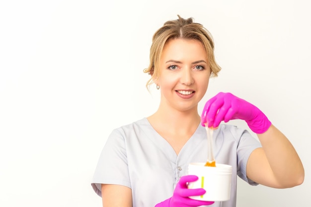 Retrato de una esteticista caucásica sosteniendo un frasco de pasta de azúcar para azucarar usando guantes rosas sobre fondo blanco