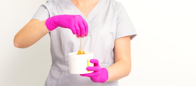 Retrato de una esteticista caucásica sosteniendo un frasco de pasta de azúcar para azucarar usando guantes rosas sobre fondo blanco