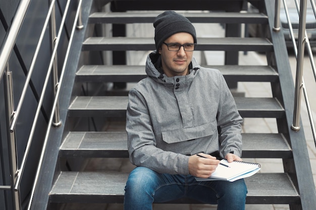 Retrato de estilo callejero de un apuesto hombre de 35 años al aire libre sentado en las escaleras y escribiendo