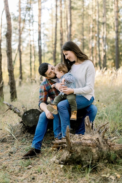 Retrato de estilo de vida al aire libre de la feliz familia caucásica, padre, madre y niño pequeño, con ropa casual elegante, sentado en un tronco en el bosque de pinos de otoño y sonriendo a la cámara