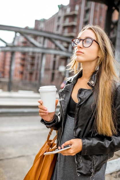 Retrato de estilo de vida de una elegante empresaria en chaqueta de cuero caminando con taza de café al aire libre en el puente de hierro