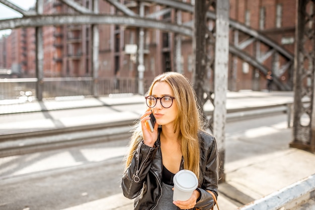 Retrato de estilo de vida de una elegante mujer de negocios en chaqueta de cuero sentada con smartphone y taza de café en el viejo puente de hierro