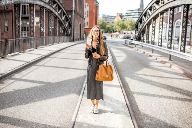 Retrato de estilo de vida de una elegante mujer de negocios de pie con una taza de café al aire libre en el viejo puente de hierro