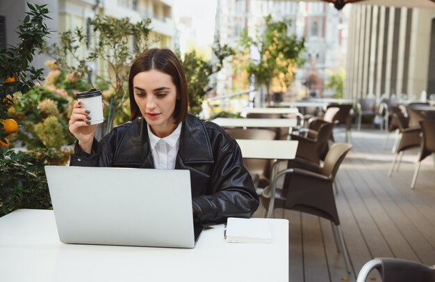 Retrato de estilo de vida de una hermosa joven confiada, persona de negocios, autónomo, desarrollador, periodista, redactor publicitario tomando café mientras trabaja en una computadora portátil, disfrutando del trabajo a distancia en el café exterior