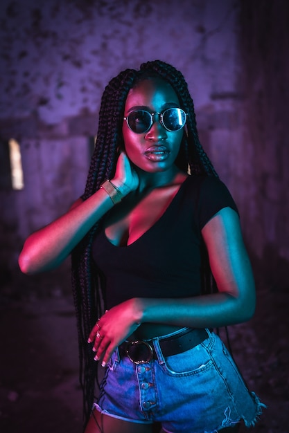 Retrato de estilo de vida de una joven mujer negra con largas trenzas gafas de sol y una camiseta negra Luces de neón rosa y azul fotografía urbana de una bailarina de trampa