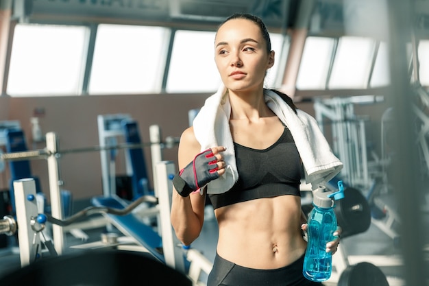 Retrato de estilo de vida de una mujer deportiva joven sonriente con una toalla blanca sobre su hombro y una botella de agua azul descansando entre ejercicios en un gimnasio