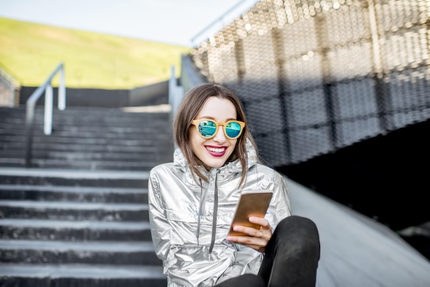 Retrato de estilo de vida de una mujer elegante en chaqueta plateada sentada con teléfono al aire libre en el entorno arquitectónico moderno