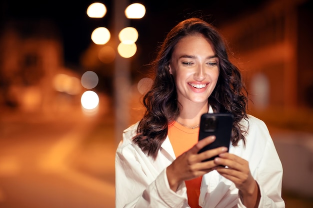 Retrato de estilo de vida de una mujer feliz caminando por la ciudad por la noche y usando el teléfono celular para enviar mensajes de texto o navegar