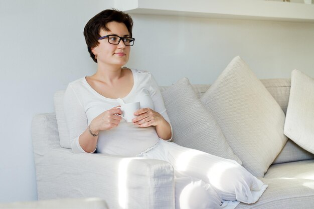 Retrato de estilo de vida de mujer feliz con falda blanca, camisa relajante, sentada. sofá. hogar acogedor. Café