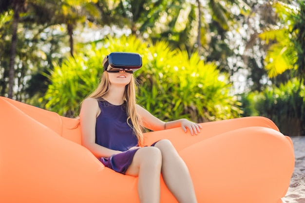 Retrato de estilo de vida de verano de una linda chica sentada en el sofá inflable naranja y usa auriculares de realidad virtual en la playa de una isla tropical Relajándose y disfrutando de la vida en una cama de aire