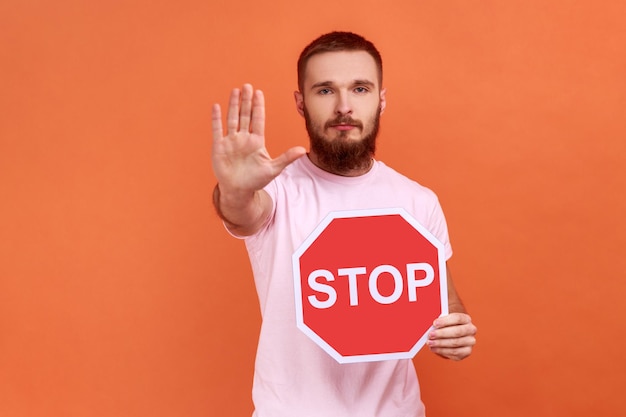 Retrato de un estricto hombre barbudo que muestra un gesto de parada y sostiene una señal de parada roja, prohibiciones y restricciones, usando una camiseta rosa. Disparo de estudio interior aislado sobre fondo naranja.