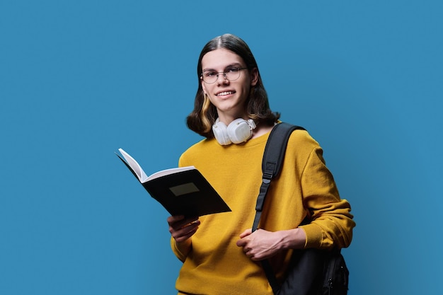 Retrato de un estudiante adolescente en fondo azul de estudio Hombre joven con gafas auriculares sosteniendo un cuaderno mirando a la cámara Escuela secundaria educación universitaria conocimiento ciencia concepto juvenil