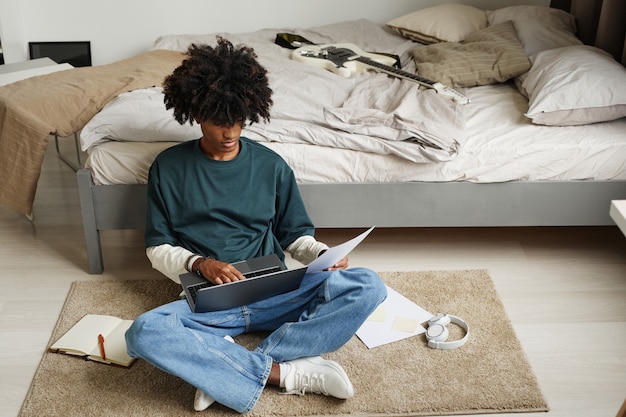 Retrato de estudiante afroamericano adolescente sentado en el piso en casa o en el dormitorio de la universidad y estudiando ...