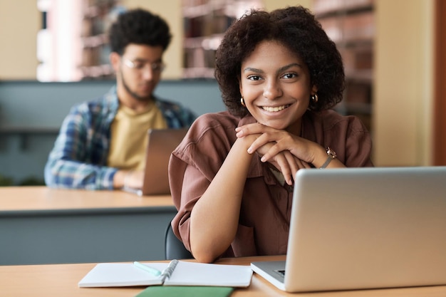 Retrato de estudiante afroamericano sonriendo a la cámara mientras se sienta en el escritorio con una computadora portátil en clase