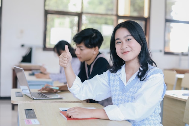Retrato de una estudiante asiática sonriente sentada mientras levanta el pulgar en el escritorio en el aula