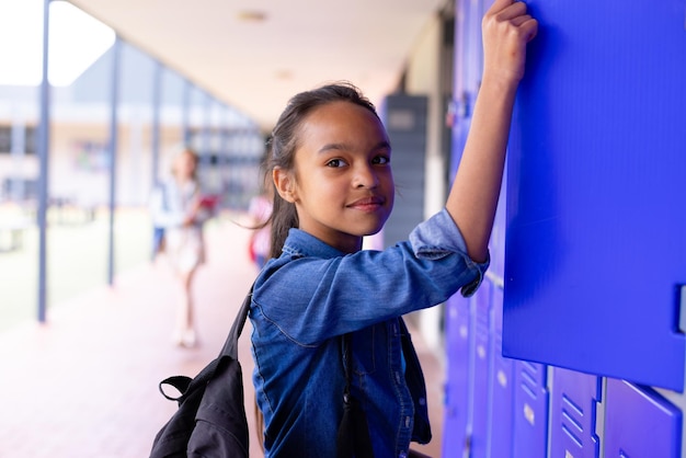 Foto retrato de una estudiante biracial feliz de pie junto a los casilleros en el pasillo de la escuela