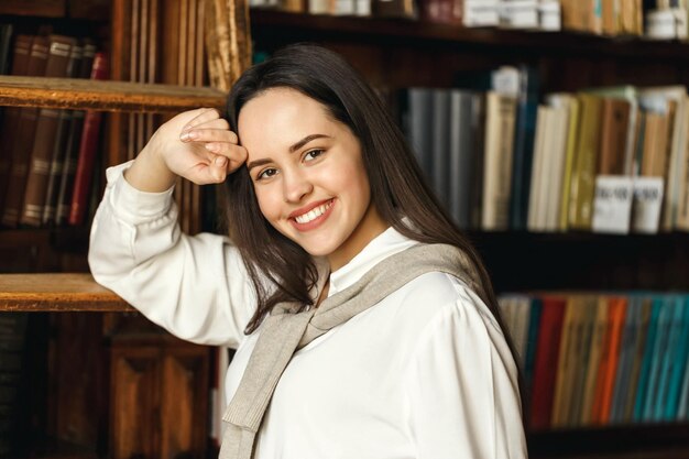 Retrato de una estudiante con camisa blanca parada y sonriendo en el fondo de las estanterías