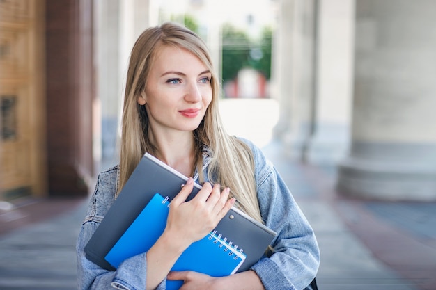 Retrato de estudiante chica caucásica sosteniendo la carpeta, cuaderno cuaderno en las manos y mirando hacia la derecha, espacio de copia