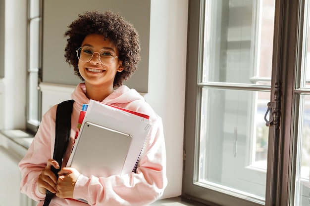 Retrato de una estudiante feliz sosteniendo libros mientras está de pie contra la ventana