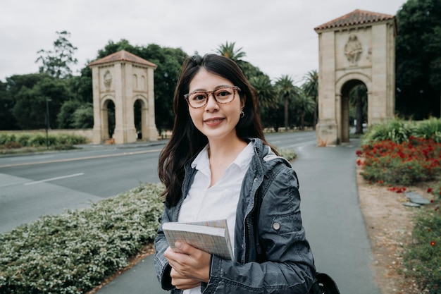 Retrato de una estudiante feliz sosteniendo libros y mirando la cámara al aire libre. joven universitaria asiática con gafas estudia en la universidad de stanford palo alto. mujer caminando por la carretera en otoño.