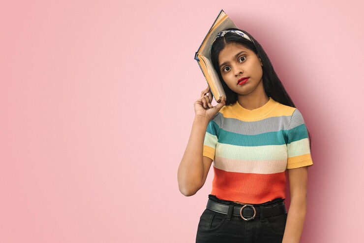 Retrato de una estudiante india asiática sorprendida con grandes ojos posando aislada sosteniendo un libro a un lado de su cabeza y mirando directamente a la cámara con espacio para copiar