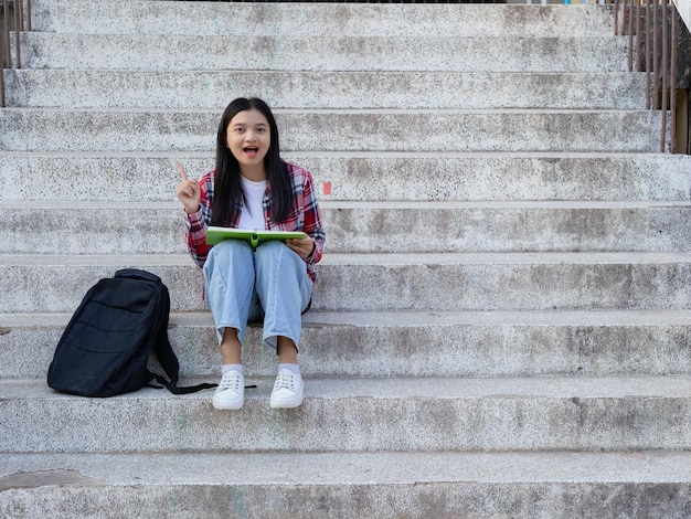 Retrato de una estudiante joven leyendo un libro y sentada en la escalera con una mochila en la escuela