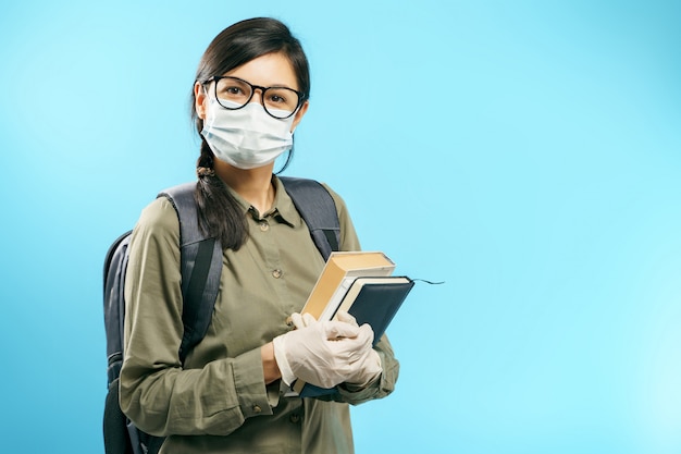 Retrato de una estudiante en una máscara de protección médica y guantes sosteniendo libros sobre un fondo azul.