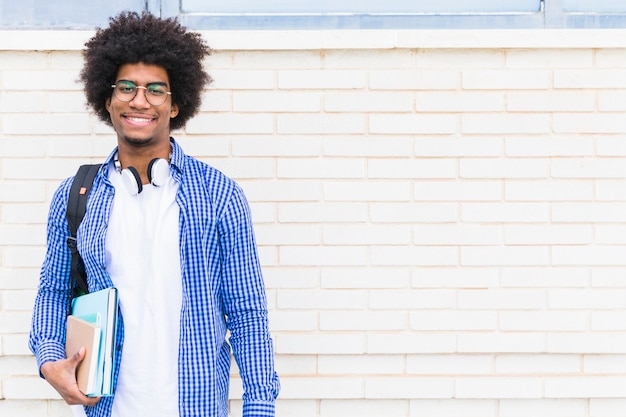 Foto retrato del estudiante masculino afroamericano sonriente que se opone a la pared de ladrillo blanca