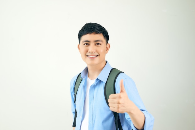 Retrato de un estudiante masculino sonriente con mochila mostrando los pulgares hacia arriba sobre la pared blanca