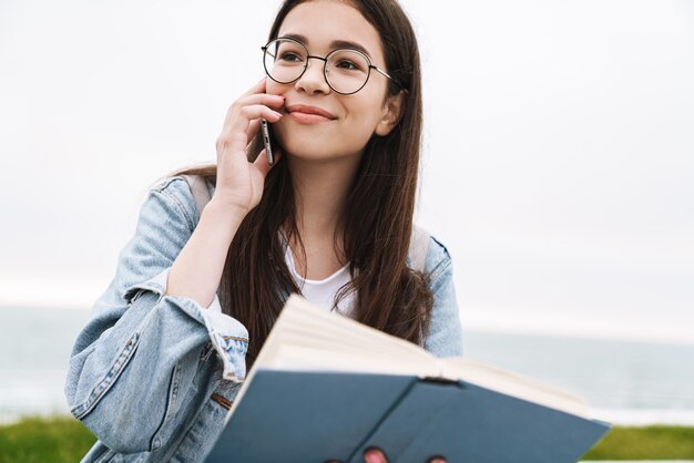 Retrato de un estudiante de mujer bonita joven emocional alegre con anteojos caminando al aire libre leyendo un libro hablando por teléfono móvil.