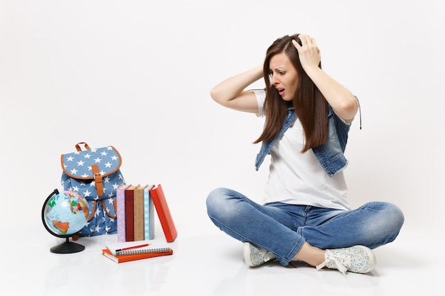 Retrato de estudiante mujer insatisfecha irritada aferrándose a la cabeza mirando hacia abajo y sentado cerca del globo, mochila, libros escolares aislados en la pared blanca