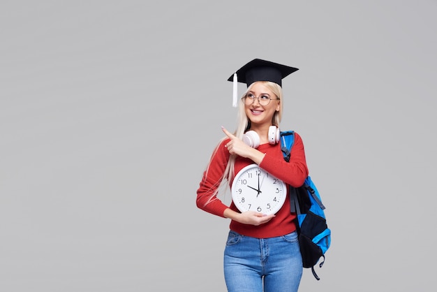 Retrato del estudiante de mujer rubio emocionado joven en casquillo graduado con la mochila que sostiene el despertador grande aislado en espacio gris. Educación en la universidad. Copiar espacio para texto