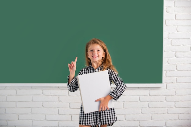 Retrato de estudiante de nerd de niña de la escuela mantenga el libro con expresión sorprendente apuntando con el dedo contra la pizarra. Educación y conocimiento de los niños. Niños estudiantes.