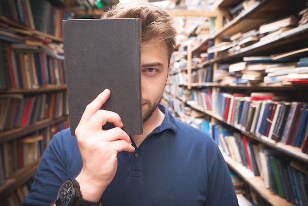 Retrato de un estudiante parado en la biblioteca, cubriéndose la cara con un libro negro