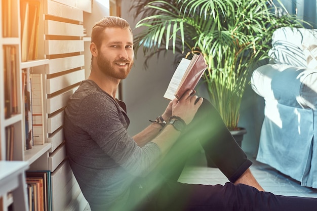 Retrato de un estudiante pelirrojo elegante y sonriente vestido con ropa informal, sentado en el suelo de una casa y sostiene un libro, mirando una cámara.