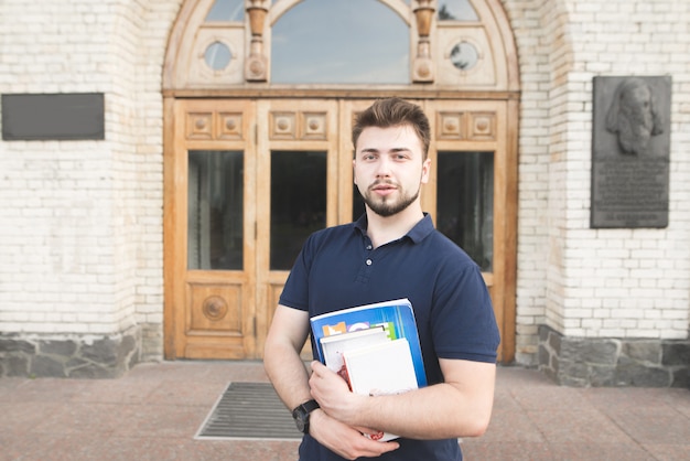 Retrato de un estudiante de pie con libros y cuadernos en la entrada de la universidad y mirando a la cámara. Hombre con barba en el fondo de una puerta de la universidad.